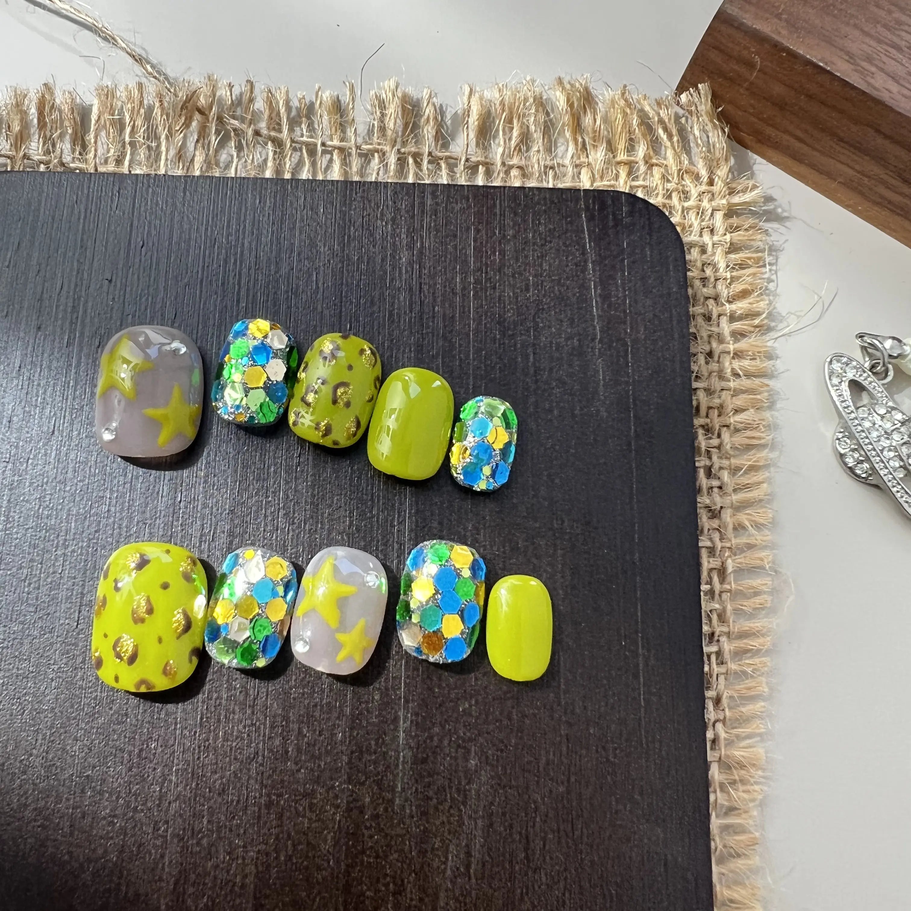 A set of neon green and glittery press on toenails with star, leopard, and chunky sequin designs, displayed on a black board.