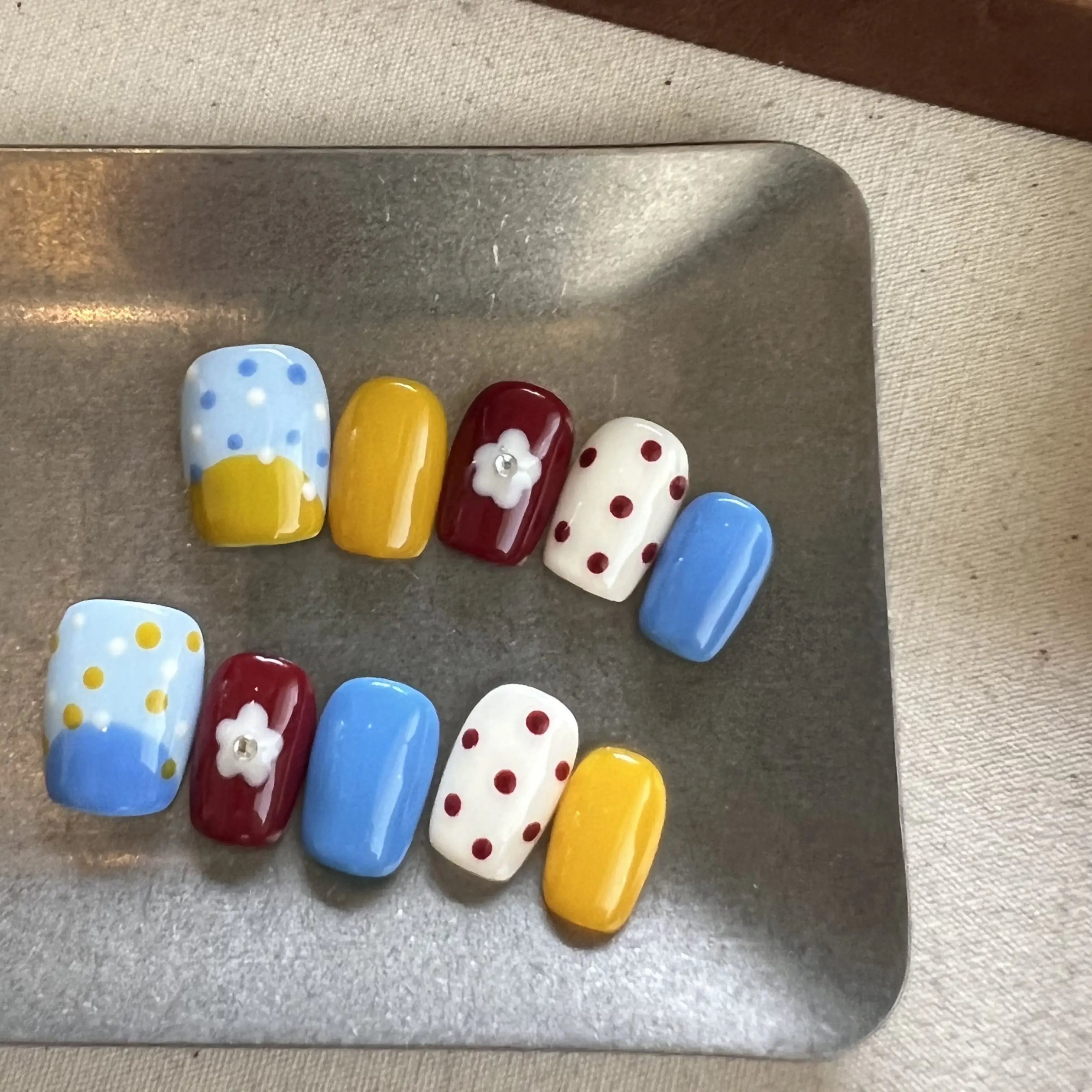 Hands with press on nails in blue, yellow, red, and white, featuring polka dots, color blocking, and 3D daisy flower accents.