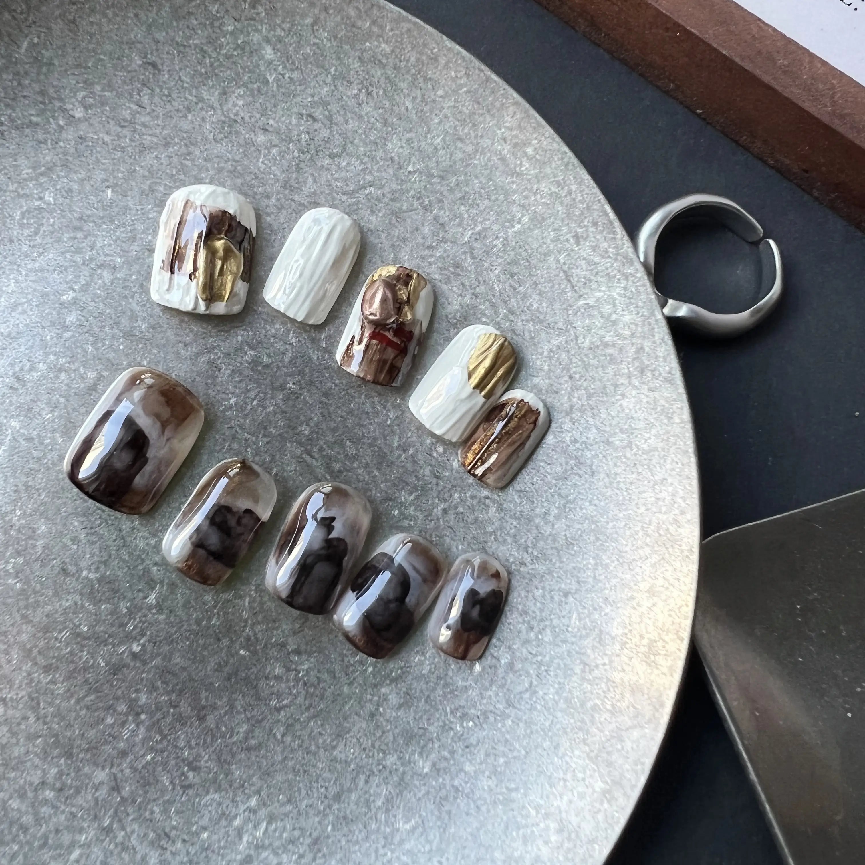 A set of press on nails in marble mocha, creamy white, and gold foil, featuring cable-knit texture and glossy finish, displayed on a card.