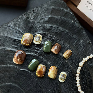 A set of press on nails with marble brown, olive green, cream, and 3D gemstone gold accents, styled on a black textured background.