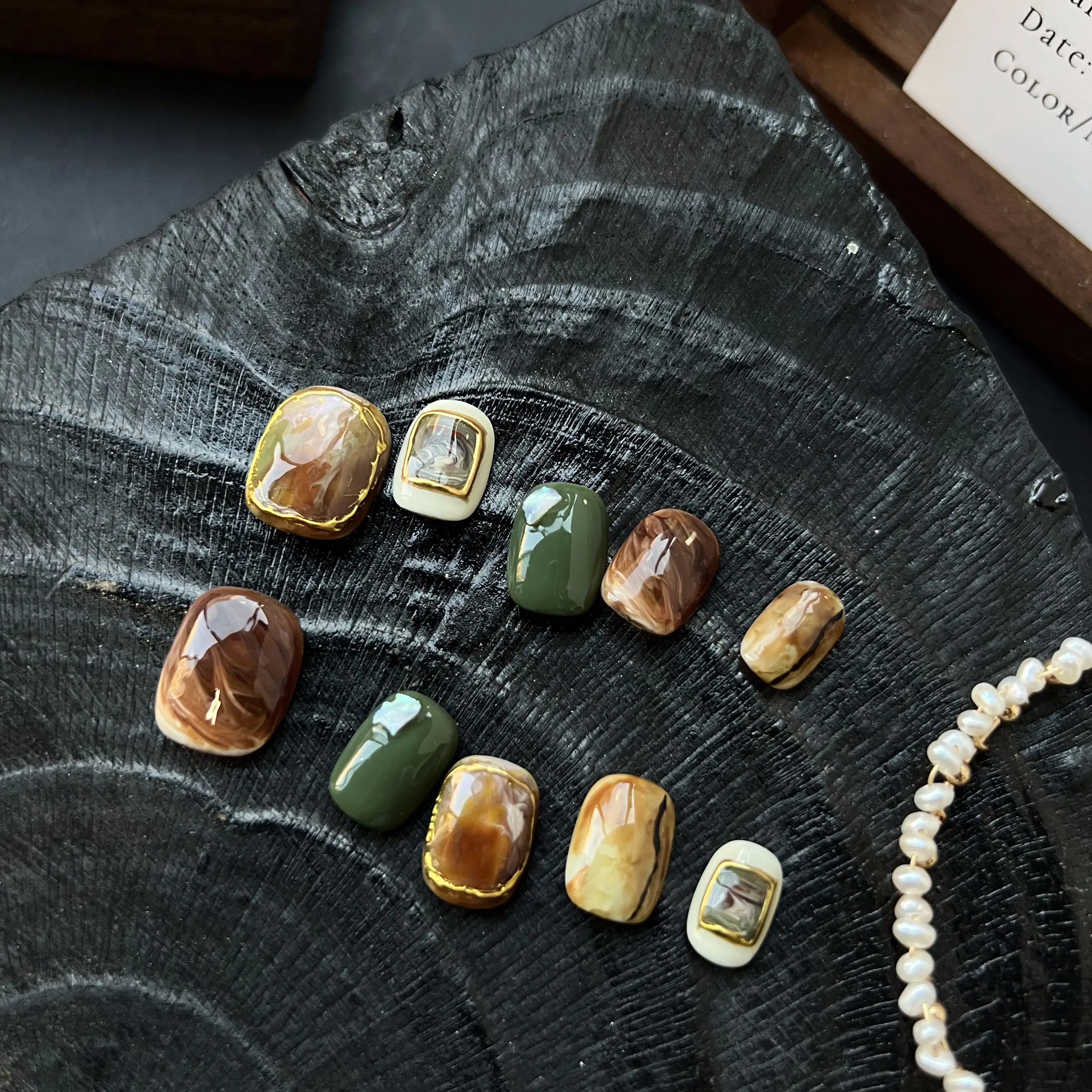 A set of press on nails with marble brown, olive green, cream, and 3D gemstone gold accents, styled on a black textured background.