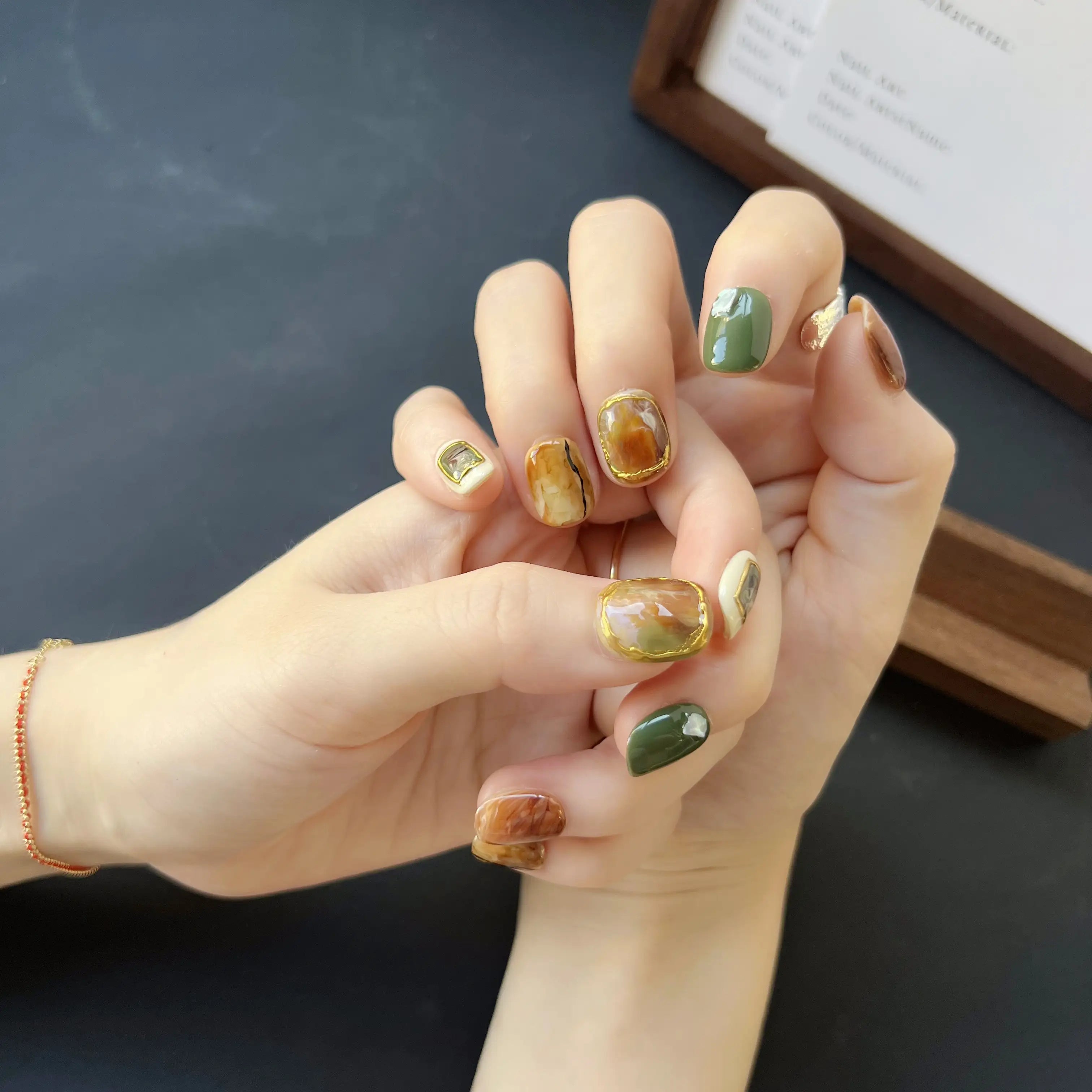 A set of press on nails with marble brown, olive green, cream, and 3D gemstone gold accents, styled on a black textured background.