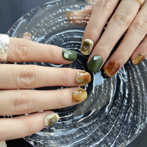 A set of press on nails with marble brown, olive green, cream, and 3D gemstone gold accents, styled on a black textured background.