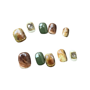 A set of press on nails with marble brown, olive green, cream, and 3D gemstone gold accents, styled on a black textured background.