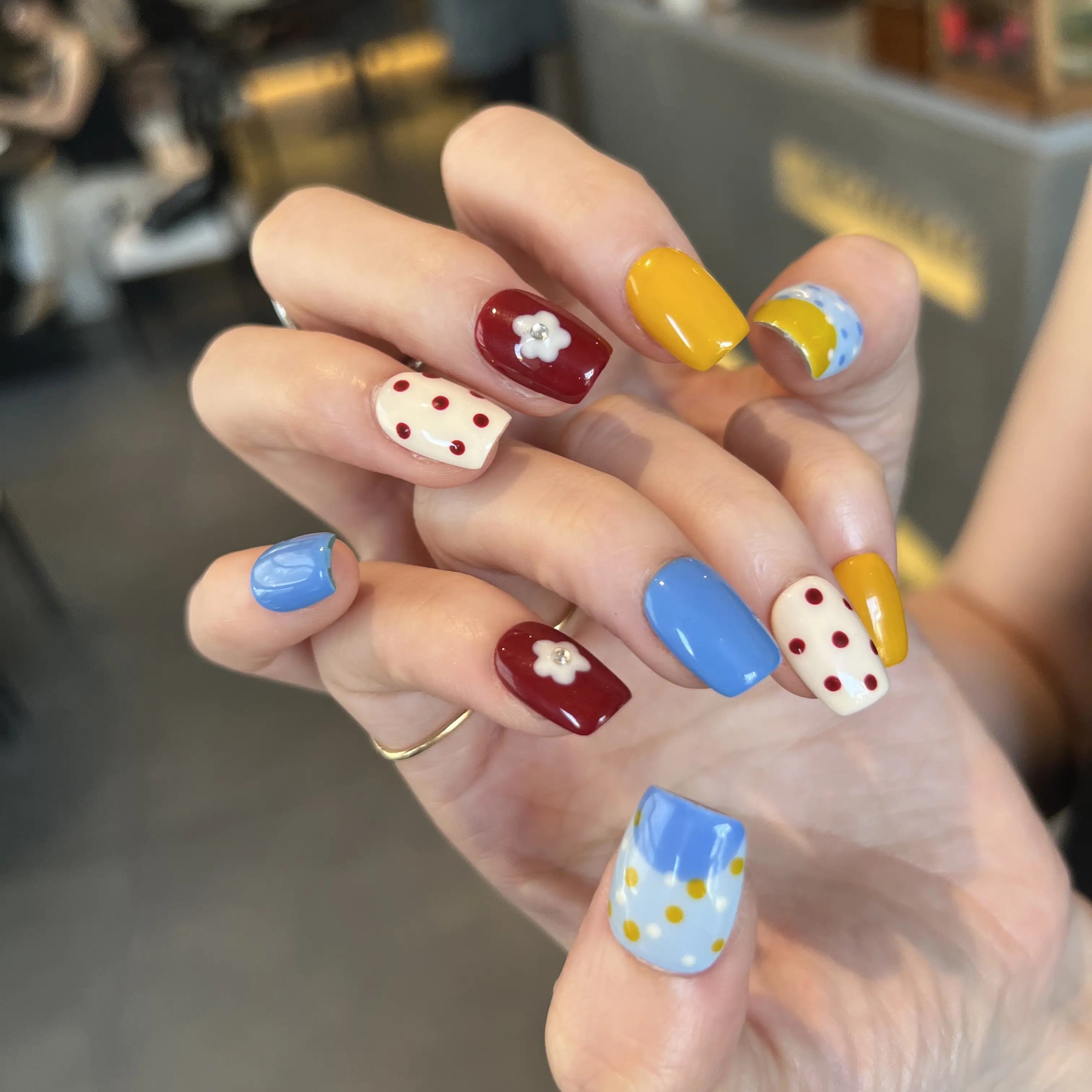 Hands with press on nails in blue, yellow, red, and white, featuring polka dots, color blocking, and 3D daisy flower accents.