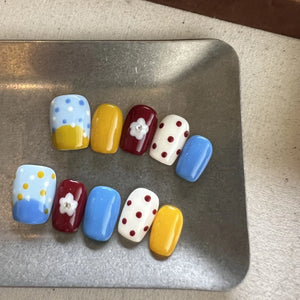Hands with press on nails in blue, yellow, red, and white, featuring polka dots, color blocking, and 3D daisy flower accents.