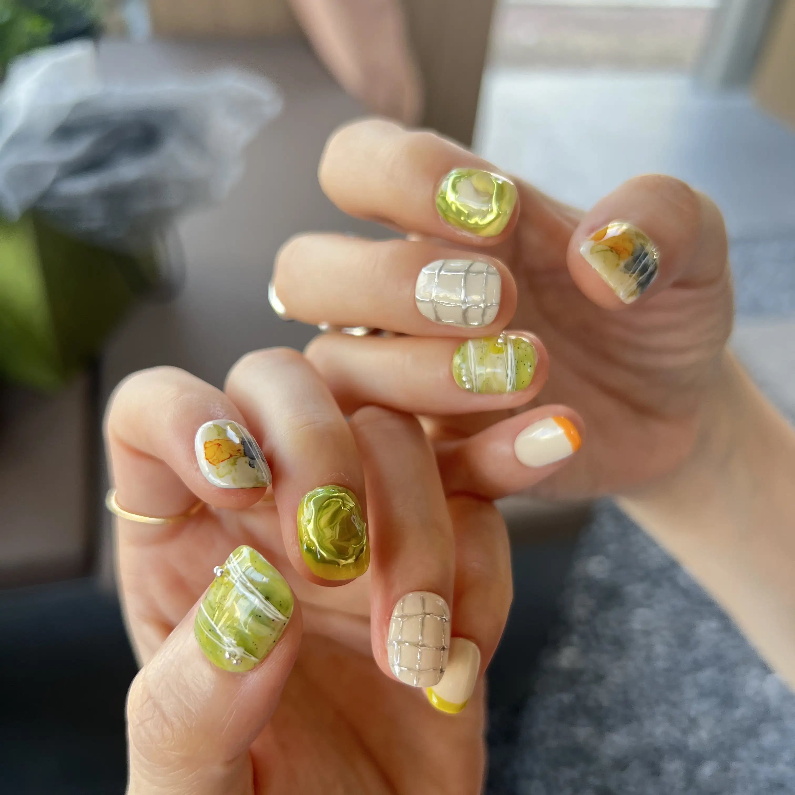 Hands with press on nails in green, white, and yellow, featuring abstract marble swirls, 3D accents, and modern grid line designs.