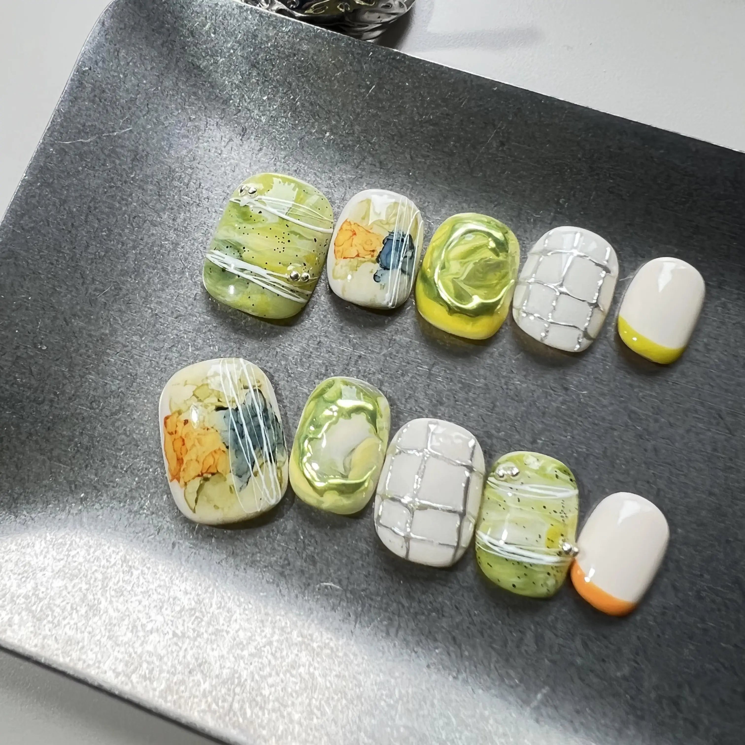 Hands with press on nails in green, white, and yellow, featuring abstract marble swirls, 3D accents, and modern grid line designs.