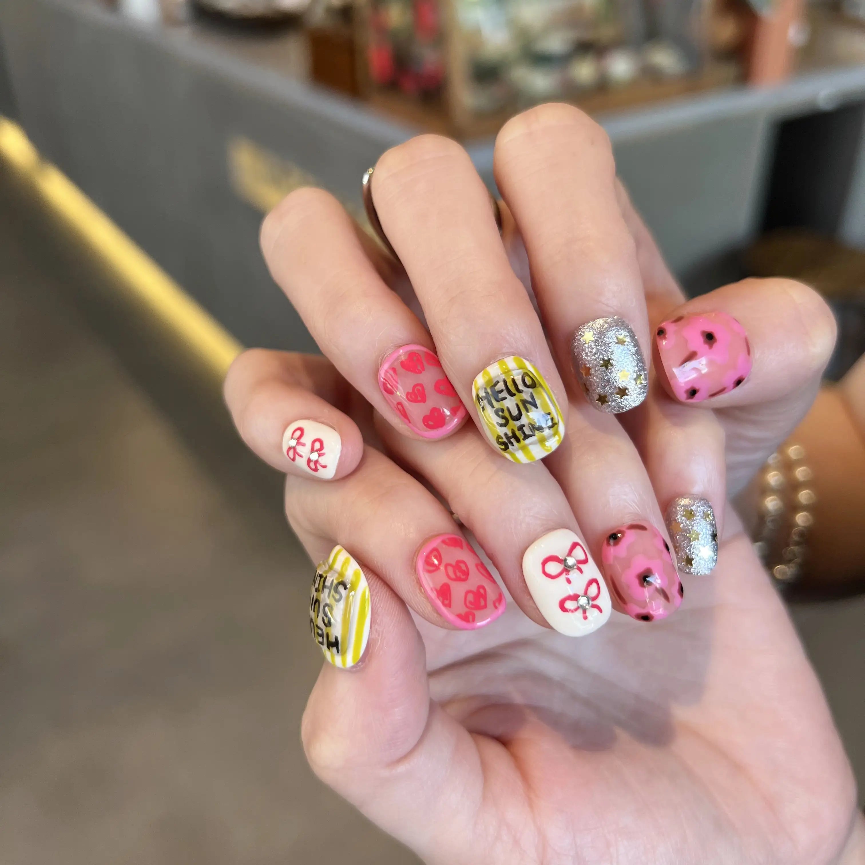 Hands with press on nails in pink, yellow, and silver glitter, featuring hand-drawn hearts, bows, and Hello Sunshine text.