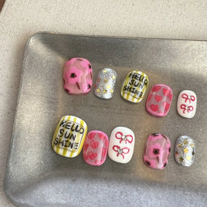 Hands with press on nails in pink, yellow, and silver glitter, featuring hand-drawn hearts, bows, and Hello Sunshine text.