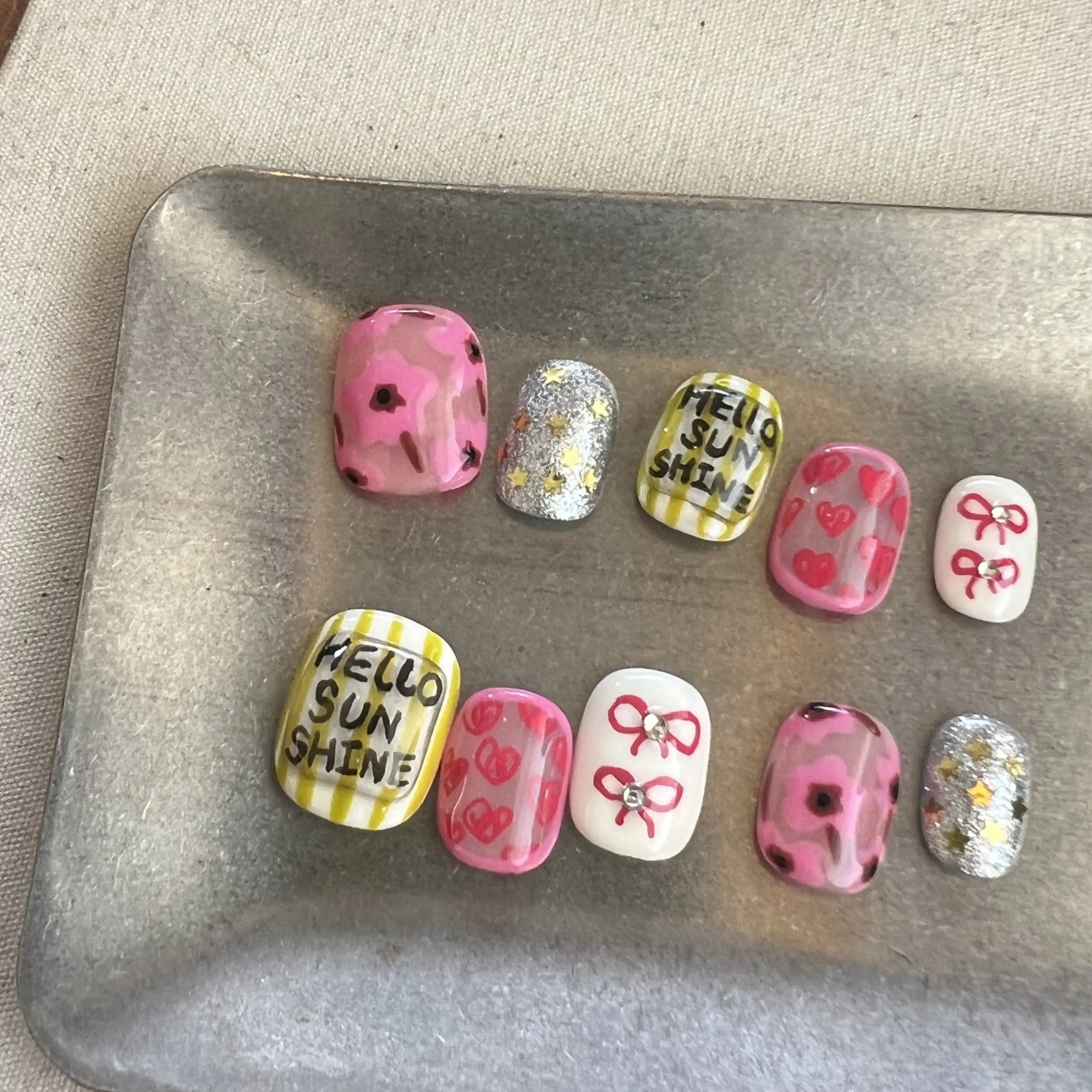 Hands with press on nails in pink, yellow, and silver glitter, featuring hand-drawn hearts, bows, and Hello Sunshine text.